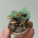 Small sculpture with clay fish and plants held by a hand against a light gray background