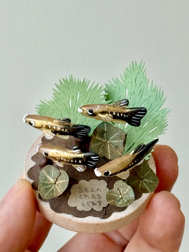 Small sculpture with clay fish and plants held by a hand against a light gray background