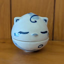 Ceramic sculpture of a blue cat jar, with a shallow bowl for the bottom and a cat head for the lid. The cat has simply drawn patterning, ears and closed eyes with thick eyeliner.
