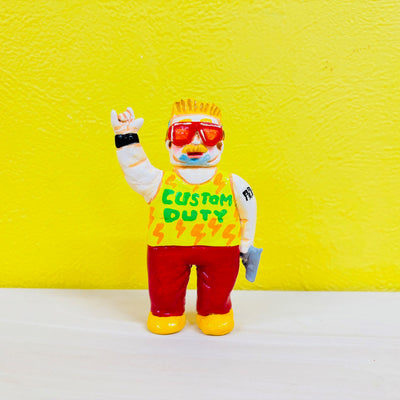 Clay sculpture of a man with short, spiky blonde hair and big red sunglasses. He holds a pistol in one hand and raises his other in a "rock on" hand shape. He wears a tank top that says "custom duty"