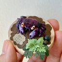 Hand holding a sculpture on a wooden disc of a purple vampire crab with paper greenery and clay rocks.