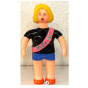 Vintage-style figurine with yellow hair, black shirt, blue shorts, and pink sash against a textured white wall.