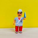 Clay sculpture of a sushi chef with thick eyebrows, he wears a white sushi chef hat and a gray kimono coat. One of his arms is a shark fin.