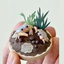 Hand holding a sculpture on a wooden disc of 2 crabs, one is a baby, with paper greenery and clay rocks.