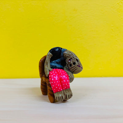 Clay sculpture of a very old person, dramatized to be depicted with brown wrinkly skin, recessed eyes and nose and nearly folded over. They sit in a chair with a black shirt and bright pink pajama pants.