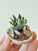 Miniature diorama with crabs and plants held in a hand against a light background
