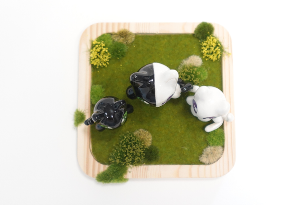 Three cat shaped figurines, with cute, large anime style eyes standing on a wooden tray with fake grass. They all have 2 tails. Seen from overhead.
