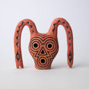 Orange ceramic clay sculpture of a face with circular holes for eyes and mouth and a triangular hole for a nose. Coming out of the top of its head like bunny ears are 2 snake like creatures, with squiggle patterning and one eye each.