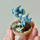 Small sculpture of 2 blue patterned angel fish, facing to the left on a wooden base with stones and paper plants.