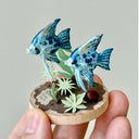 Small sculpture of 2 blue patterned angel fish, facing to the left on a wooden base with stones and paper plants.