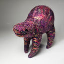 A handcrafted felted wool animal figurine, shaped like a small four-legged creature with a textured, mottled purple, pink, and orange coloration, set against a neutral gray background.