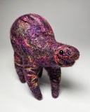 A handcrafted felted wool animal figurine, shaped like a small four-legged creature with a textured, fuzzy surface in marled purple, pink, and green tones, set against a neutral gray background.