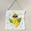 A square white fabric wall hanging with a wooden dowel and green hanging cord, featuring embroidered artwork of a leafy yellow creature with pink and orange details, on a neutral beige background.