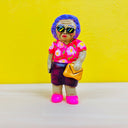 Clay sculpture of an older woman with greenish brown skin, sunglasses and short permed purple hair. She wears a purple and pink floral shirt, purple skirt and holds a purse.