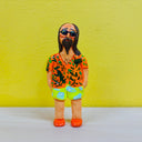 Clay sculpture of a man with long brown hair and a long beard. He is dressed in beach wear and stands with his head tilted slightly to the side. His short is orange with green patterning, his shorts are green and blue and his shoes are orange