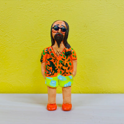 Clay sculpture of a man with long brown hair and a long beard. He is dressed in beach wear and stands with his head tilted slightly to the side. His short is orange with green patterning, his shorts are green and blue and his shoes are orange