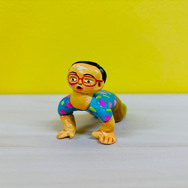 Clay sculpture of a yellow man with glasses and a surprised expression. He is kneeling on the ground with his hands touching the floor and his knees are on the ground as well.
