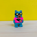 Clay sculpture of a blue cat, standing on 2 legs like a human. It wears sunglasses, a bright pink shirt with a graphic of an uncut sushi roll and has an open mouth expression.