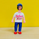 Clay sculpture of a woman wearing a white t-shirt that reads "California" with Japanese kanji written underneath, blue jeans and red shoes. She wears a baseball cap that says "sushi"