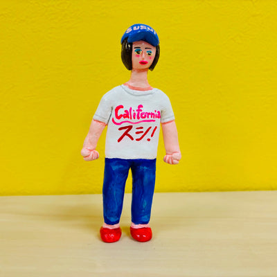 Clay sculpture of a woman wearing a white t-shirt that reads "California" with Japanese kanji written underneath, blue jeans and red shoes. She wears a baseball cap that says "sushi"