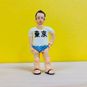 Clay sculpture of a man wearing a white t-shirt, a blue striped bathing suit briefs and sandals. He has one hand on his hip and his penis peeks out of the bathing suit.