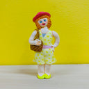 Clay sculpture of a smiling woman with short orange hair with bangs, She wears a red hat and yellow sun dress. She has one hand on her hip and the other holds her purse, over her shoulder.