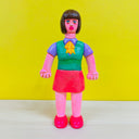 Clay sculpture of a pink woman, dressed like an office worker in bright colors - a pink skirt, purple button up, green vest  and yellow and orange polka dot handkerchief. 
