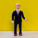 Clay sculpture of a blonde business man with short hair and a dark blue suit, pink shirt and gray tie. He holds a chunky cigarette.