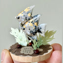 Small sculpture of 2 gray angel fish with black, white and gold spots, facing to the left on a wooden base with stones and paper plants.