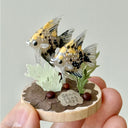 Small sculpture of 2 gray angel fish with black, white and gold spots, facing to the left on a wooden base with stones and paper plants.
