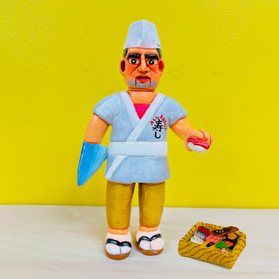 Clay sculpture of a sushi chef with thick eyebrows, he wears a white sushi chef hat and a gray kimono coat. One of his arms is a shark fin. He holds a piece of sushi in the other hand.