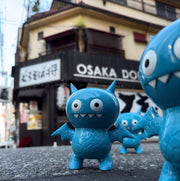 Figurine of Uglydoll Ice-Bat, with wide eyes and fanged teeth. It stands in the street with 2 more of the same figures around it. It has horns, wings and scaled body.