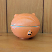 Ceramic sculpture of an orange cat jar, with a shallow bowl for the bottom and a cat head for the lid. The cat has simply drawn patterning, ears and closed eyes with thick eyeliner.