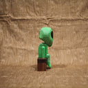 Whittled wooden sculpture of a green alien with a large head and black eyes. It has a slight frown and sits on a bench.