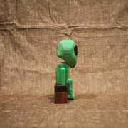 Whittled wooden sculpture of a green alien with a large head and black eyes. It has a slight frown and sits on a bench.