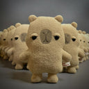 A row of plush capybara toys in a warm beige color, arranged in a diagonal line on a gray surface with a gray background. The front-most plush is in sharp focus, with the others softly blurred in the background.