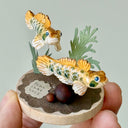 Small sculpture of 2 orange gobi fish with green spots, facing to the right on a wooden base with stones and paper plants.