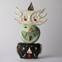 Sculptural piece consisting of 3 head like elements, stacked atop one another like a totem pole. The bottom is a black head akin to a boar, middle is a green head akin to a snake, and the top is a white head with pointed elements and red eyes possibly akin to a bat.