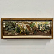 Diorama style mixed media sculpture in a long rectangular glass box of a forest floor scene, with 2 frogs dueling in the center. All around them are various cut paper leaves and plants.