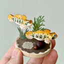 Small sculpture of 2 orange gobi fish with green spots, facing to the right on a wooden base with stones and paper plants.