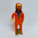 Clay sculpture of a man with a receding hairline, long brown hair and a matching beard. He wears and orange suit and stands holding a sticky note and a large pickle between his legs.