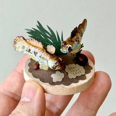 Two small fish figurines on a circular wooden base held in a hand with a plain background