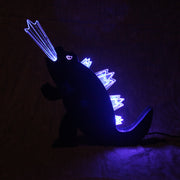 Die cut wooden silhouette of Godzilla, made into a lamp. A beam of light comes out of its mouth and it has lit up spikes along its back.