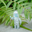Yeti-like toy figure with a skinny mushroom in its hand in front of green ferns
