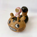 Ceramic sculpture of a cat like creature with a wide toothed grin and a large tail, without any visible limbs. A small character sits atop its head, dressed in a pink body suit.