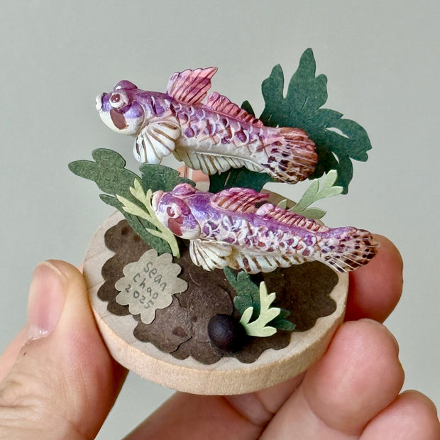Hand holding a small wooden box with paper fish and plants on a gray background