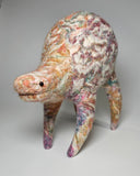 A handcrafted felted wool animal figurine with a textured, multi-colored pattern of cream, pink, orange, green, and purple, standing on a neutral gray background.