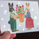 Illustration of two bunnies with a basket of flowers on a card held by a hand.