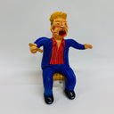 Clay sculpture of a caricature of Donald Trump, with a blown off blonde wig, a large open mouth and a blue suit with red shirt. He holds a cigar.