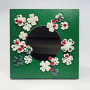 A circular mirror attached to a green panel surrounded by white ceramic flowers with red ladybugs and green accents, set against a plain light gray background.